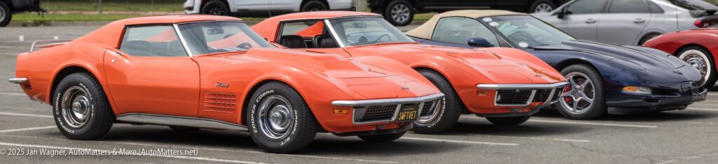 Three classic Chevrolet Corvette cars, two orange and one dark blue, are parked in a row in an outdoor parking lot.