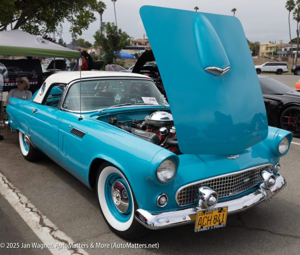 A turquoise classic car with its hood open is parked at a car show, displaying its engine. The car has whitewall tires and a California license plate.