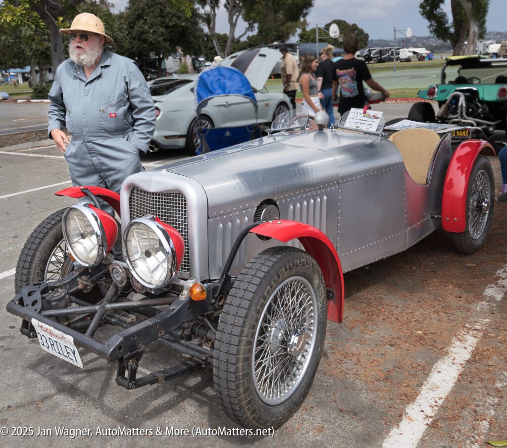 An older man in a hat stands beside a vintage silver car with red fenders at an outdoor car show. Other vehicles and people are visible in the background.