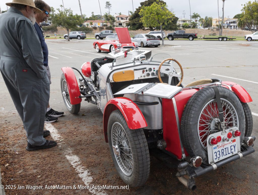 Two people stand beside a vintage red open-top car with a “33RILEY” license plate, parked in a lot with other classic cars visible in the background.