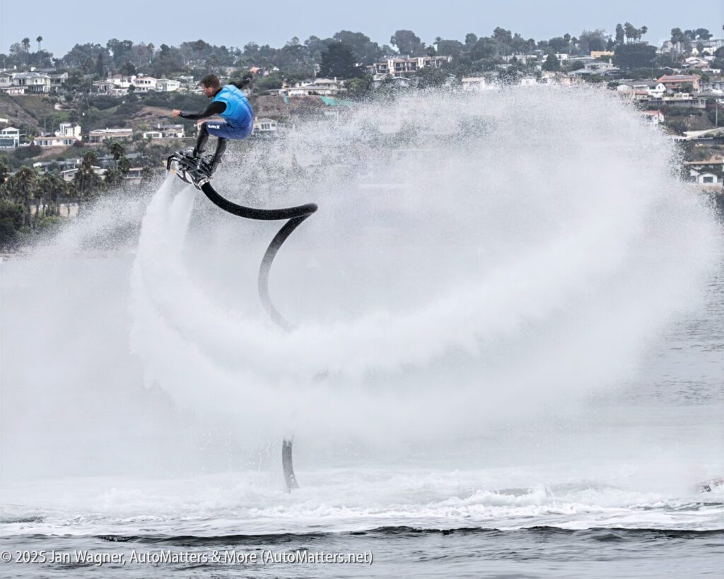A person wearing a blue shirt performs a high jump over water using a water-powered jetpack, creating a large spray, with houses visible on the distant shoreline.