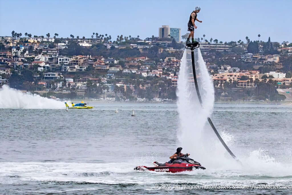 A person on a flyboard is elevated above the water, powered by a jet from a watercraft, with a cityscape and boats visible in the background.
