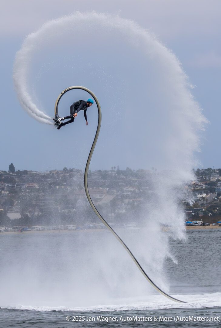 A person wearing a helmet performs a flyboard stunt over water, arcing high in the air with water spraying behind them, cityscape in the background.