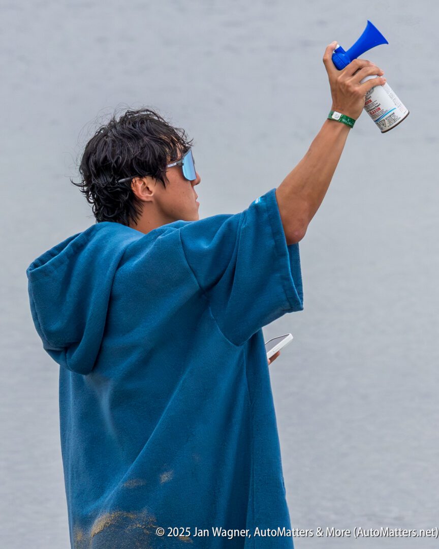 Person wearing a blue poncho holds up a blue air horn, facing away from the camera, with a body of water in the background.