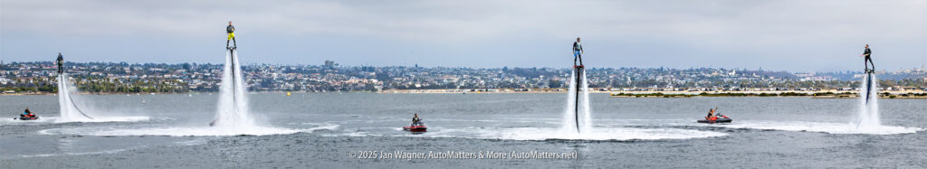 Six people are using water-powered jetpacks above a bay, each connected to a jet ski, with a cityscape in the background.