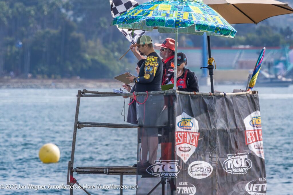 Three people stand on a judging platform by the water, shaded by umbrellas, overseeing a water event. A checkered flag and sponsor banners are visible.