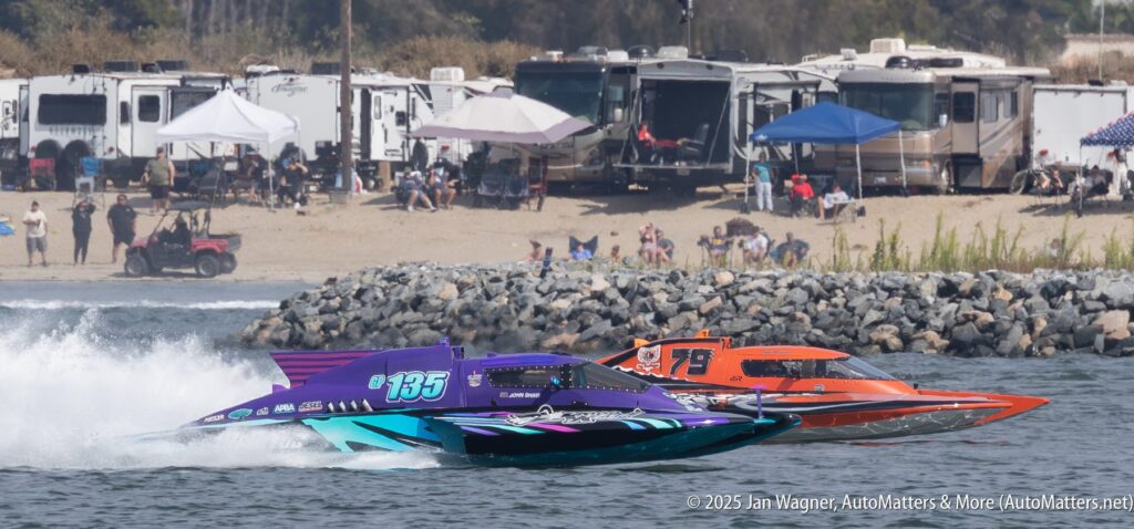 Two powerboats race side by side on the water near a rocky shoreline, with RVs, tents, and spectators visible in the background.