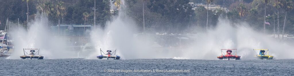 Four speedboats race across the water, creating large sprays behind them, with trees and blurred buildings in the background.