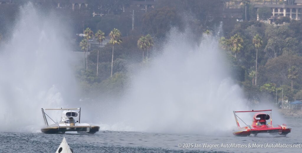 Two hydroplane boats race on water, creating large sprays behind them; trees and buildings are visible in the background.