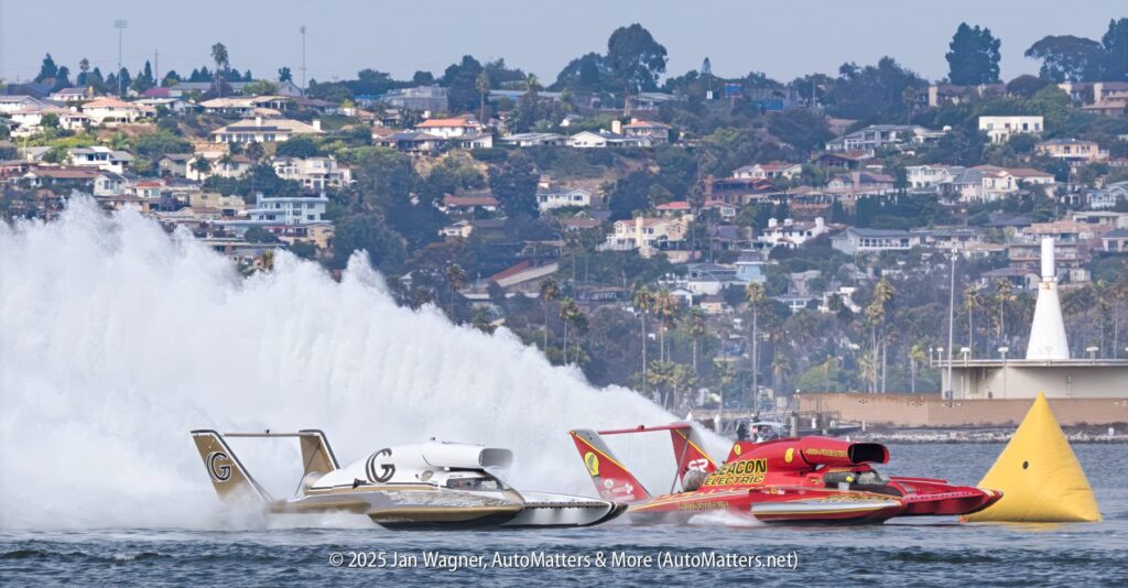 Two hydroplanes race side by side on a body of water, creating large spray, with houses and trees on a hillside in the background.