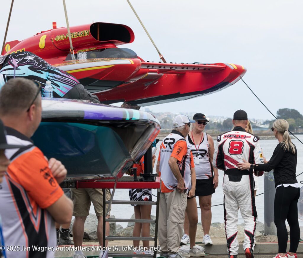 People gather near two speedboats, one red and one blue, as a man in a racing suit labeled "8" speaks with others by the waterfront.