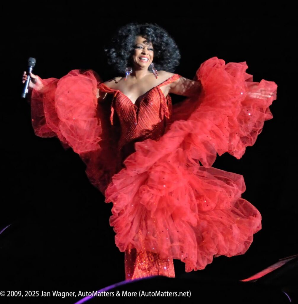 A performer in a sparkling red dress with voluminous ruffles holds a microphone and smiles on stage under bright lights.