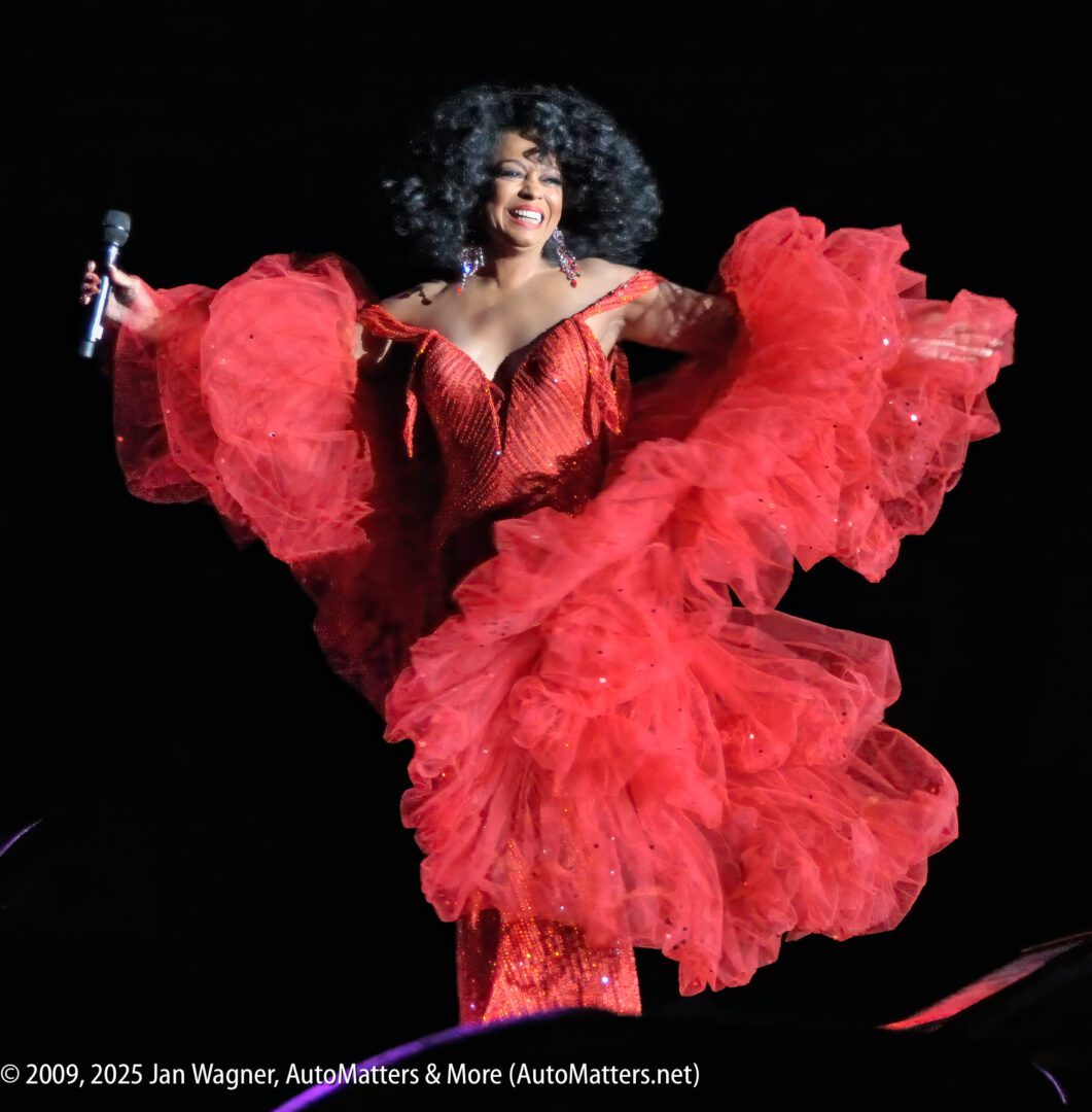 A performer in a sparkling red dress with voluminous ruffles holds a microphone and smiles on stage under bright lights.