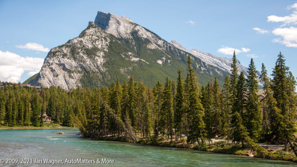 A river bordered by pine trees flows in front of a rugged, forested mountain under a partly cloudy sky.