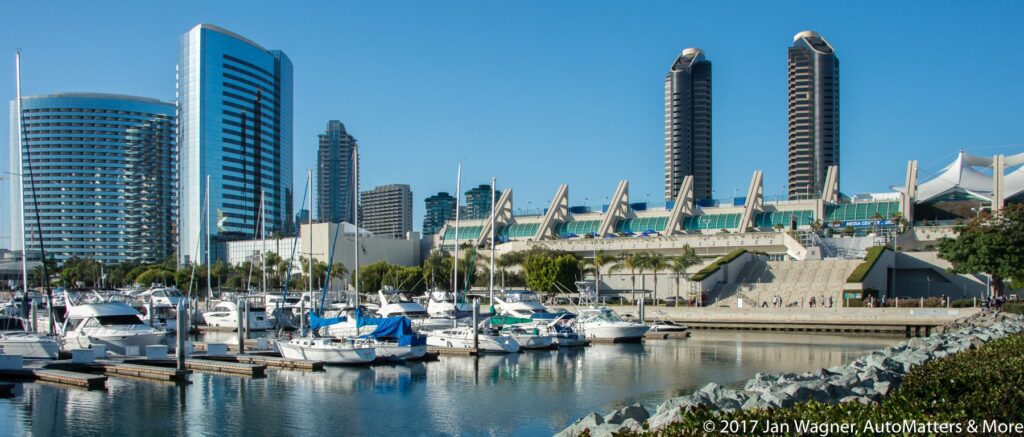 A marina with docked boats in the foreground and modern high-rise buildings and the San Diego Convention Center in the background under a clear blue sky.