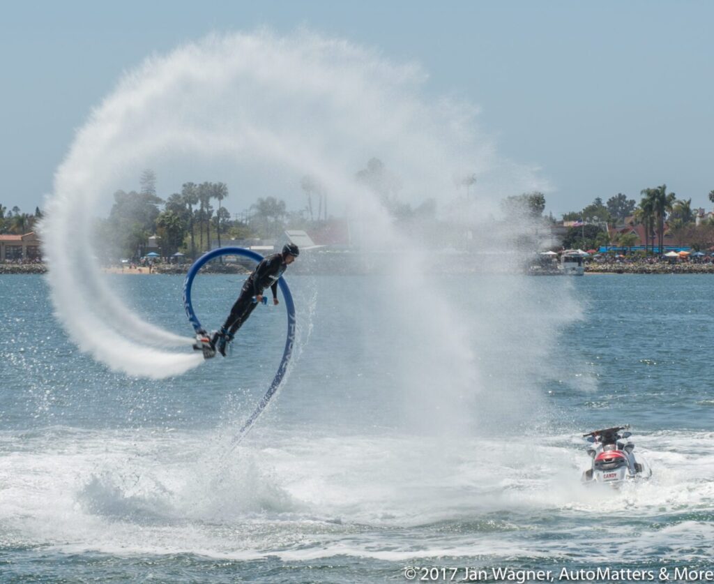 A person on a water-powered flyboard performs a high arc over the water, with a jet ski nearby and spray forming a loop in the air.