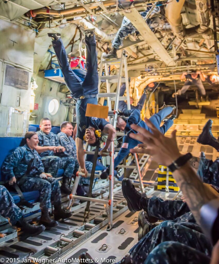A group of people in military uniforms watch as a person hangs upside down from a ladder inside an aircraft cabin.