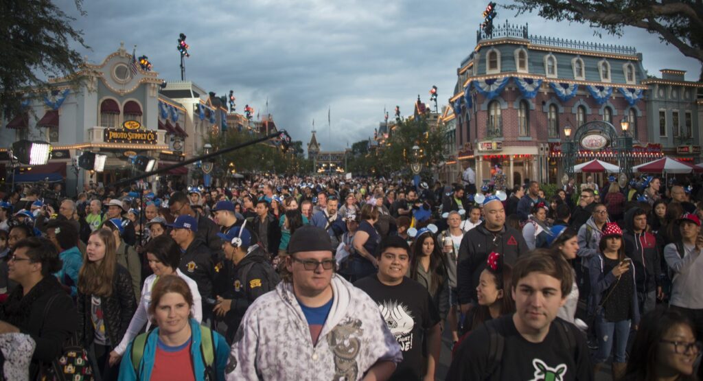 A large crowd of people walking down Main Street at Disneyland, with buildings decorated in blue and white banners under a cloudy sky.