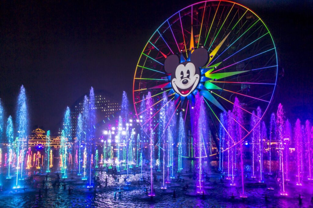 A large Ferris wheel with a Mickey Mouse face and rainbow lights is illuminated behind colorful, lit water fountains at night.