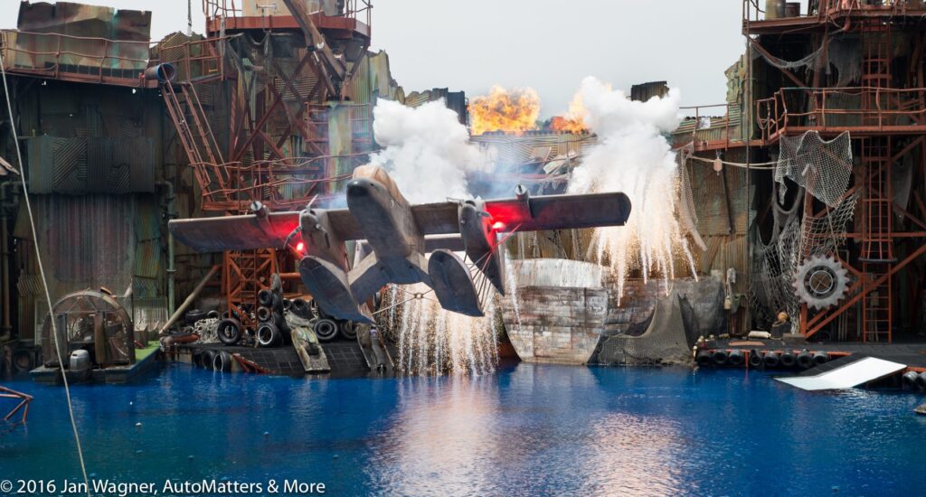 An airplane with red lights flies low over blue water as fire, smoke, and sparks erupt from a rugged industrial set in a staged action scene.
