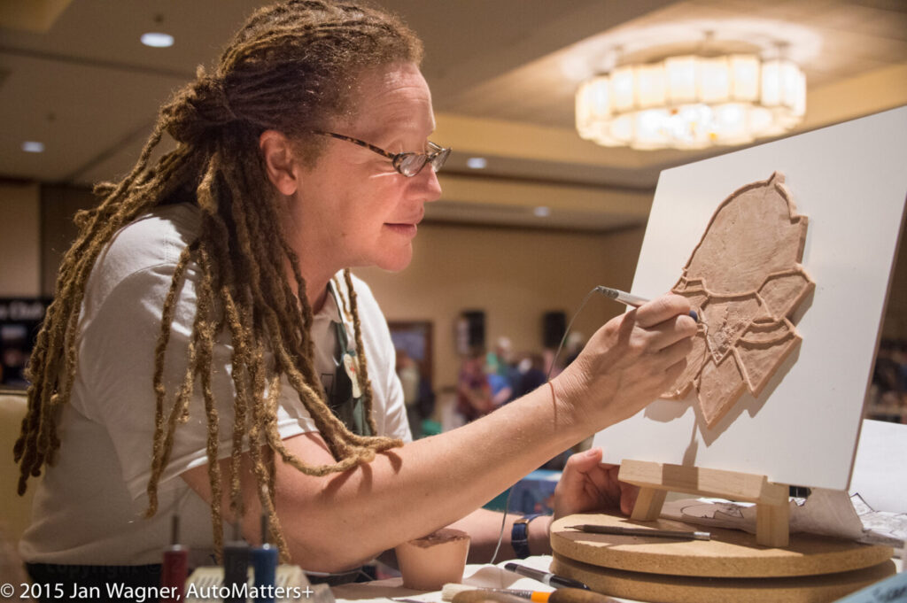 A person with dreadlocks is carving a design into a piece of leather on a worktable, using a tool, in a well-lit indoor setting.
