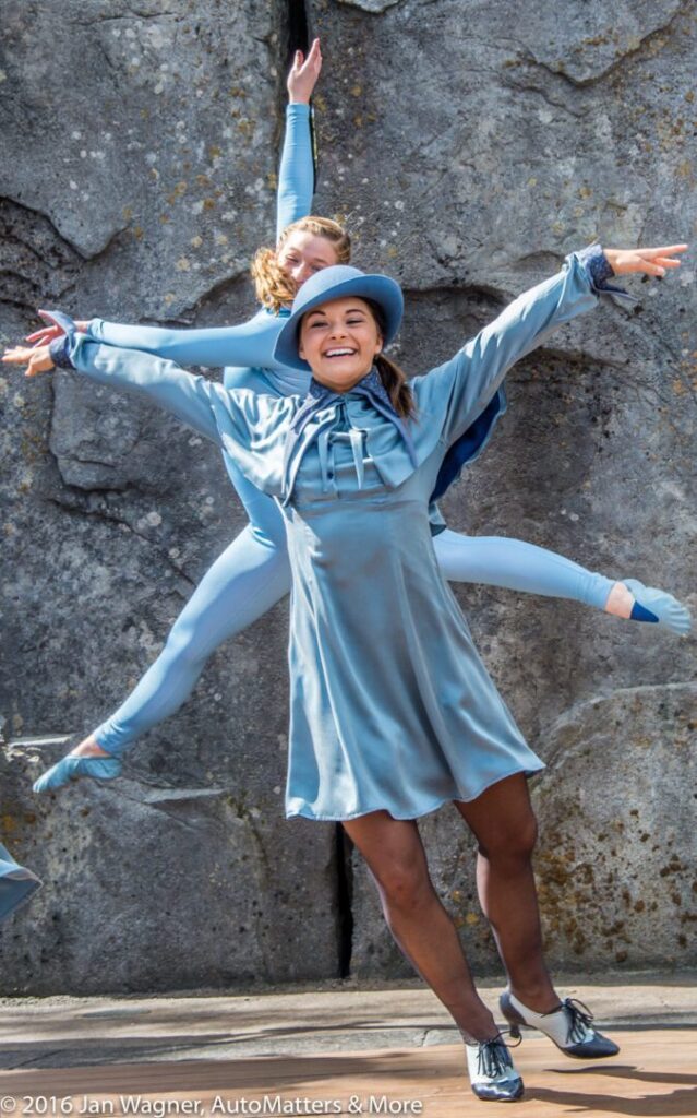 Two performers in blue costumes pose energetically, with one in a dress and hat smiling in front and another in a bodysuit leaping with arms outstretched behind her, against a stone backdrop.