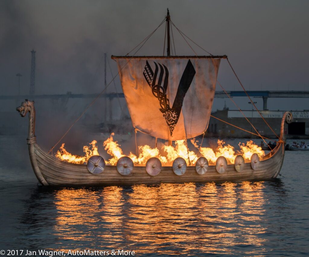 A wooden Viking-style ship with a decorated sail is engulfed in flames on the water at dusk.