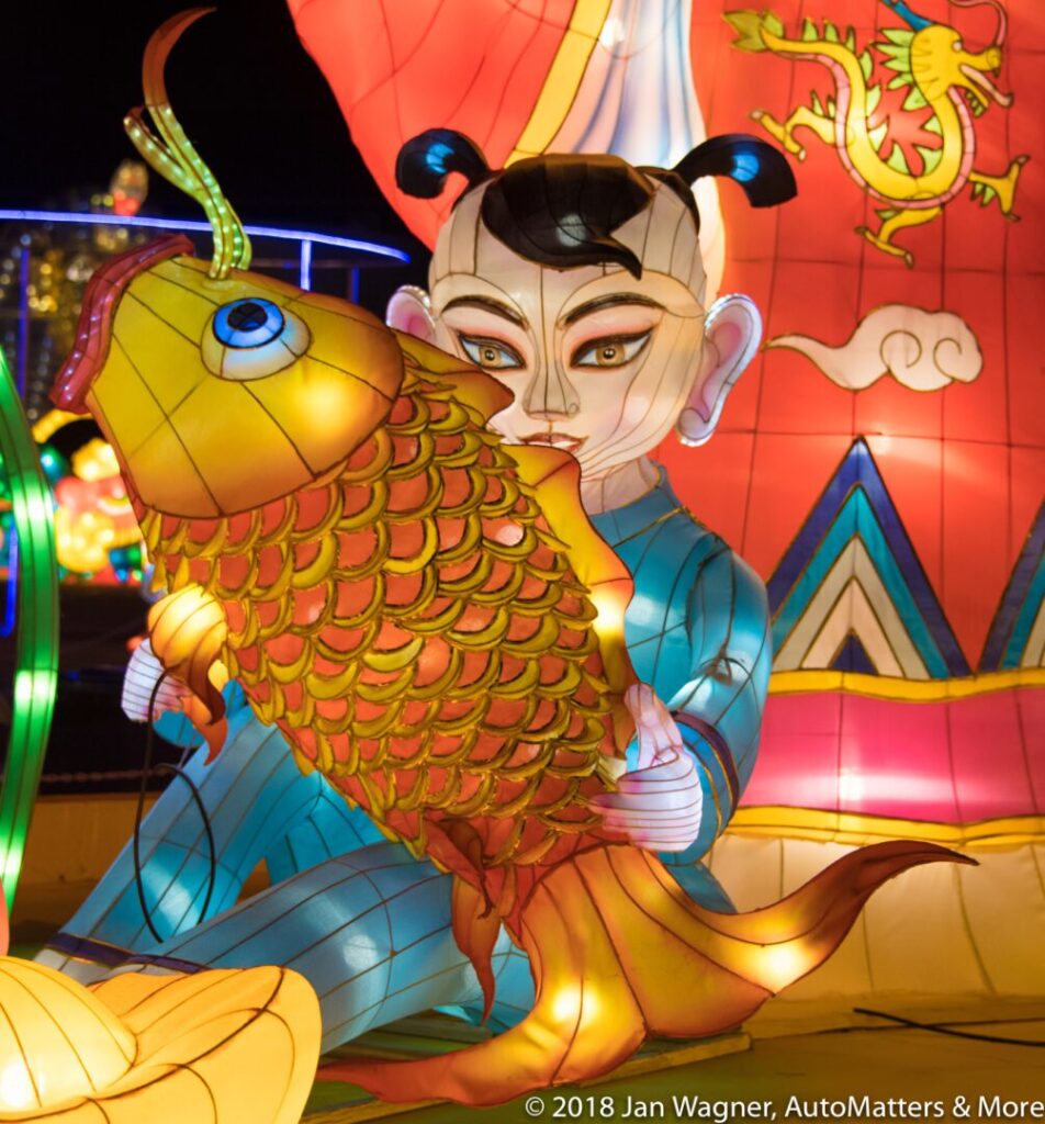 A colorful lantern display featuring a child figure holding a large fish, with a red backdrop and dragon design.