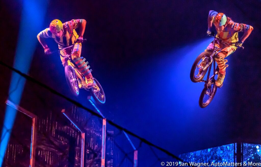 Two cyclists in colorful outfits perform mid-air tricks on BMX bikes against a dark, illuminated indoor backdrop.