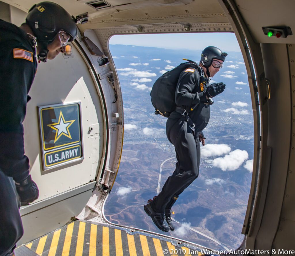 A U.S. Army paratrooper jumps out of an aircraft in mid-air while another paratrooper looks on from inside the plane.