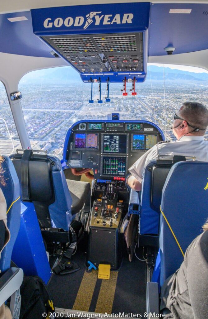 View from behind two pilots inside the Goodyear blimp cockpit, showing flight controls, instrument panels, and a cityscape through the windows.