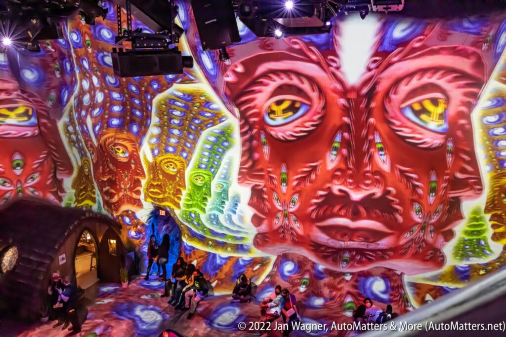 People sit in a room with large, colorful, psychedelic face patterns projected onto the walls and ceiling, creating an immersive art environment.