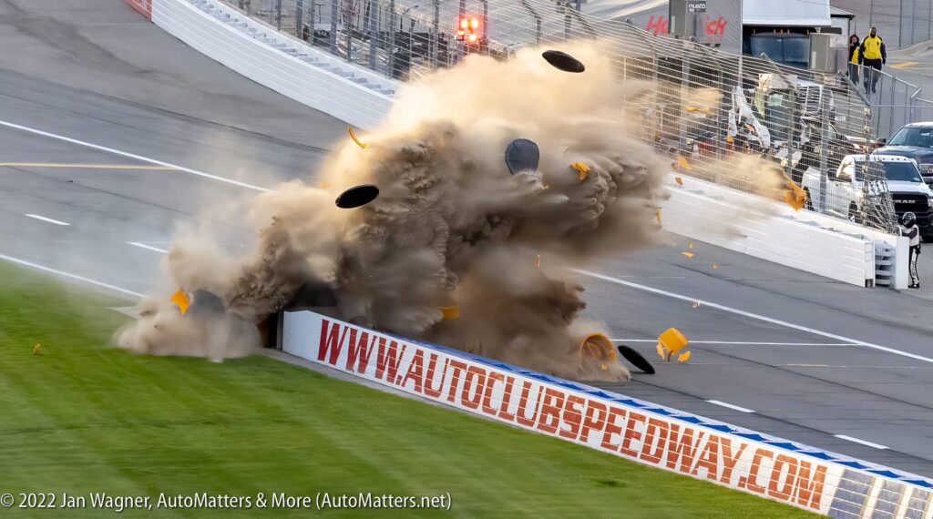 A race car crashes into a wall at high speed, causing debris, tires, and a large cloud of dust to scatter onto the racetrack.
