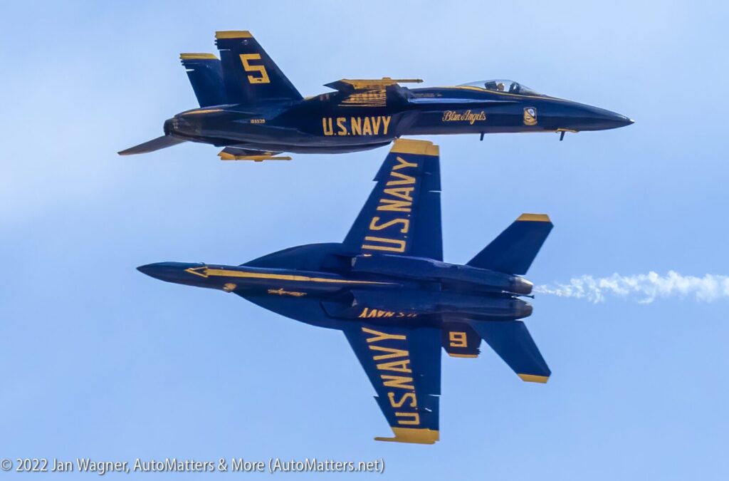 Two U.S. Navy Blue Angels F/A-18 jets perform a close, mirrored flyby maneuver against a clear sky.