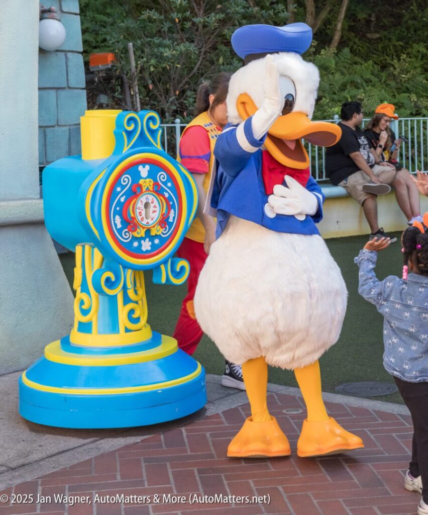 Donald Duck character in costume salutes while interacting with a child at an outdoor theme park area near a colorful blue and yellow prop.
