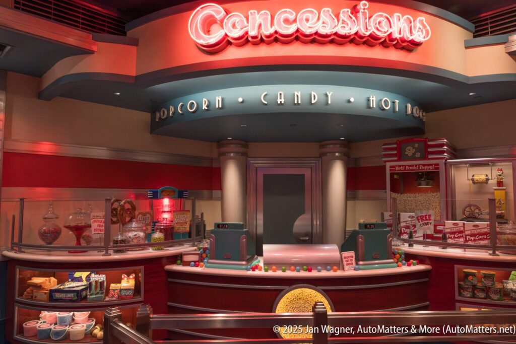 A retro-style concessions stand with popcorn, candy, hot dogs, and drinks on display, featuring bright signage and a colorful counter.