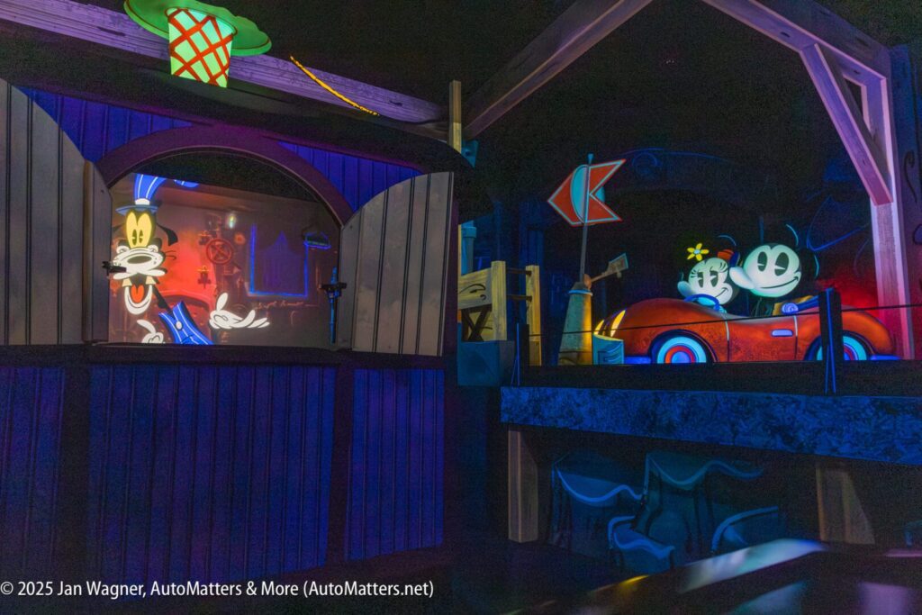 Animated figures of Goofy, Mickey Mouse, and Minnie Mouse appear in a colorful, themed indoor amusement park ride scene with vibrant lighting and props.
