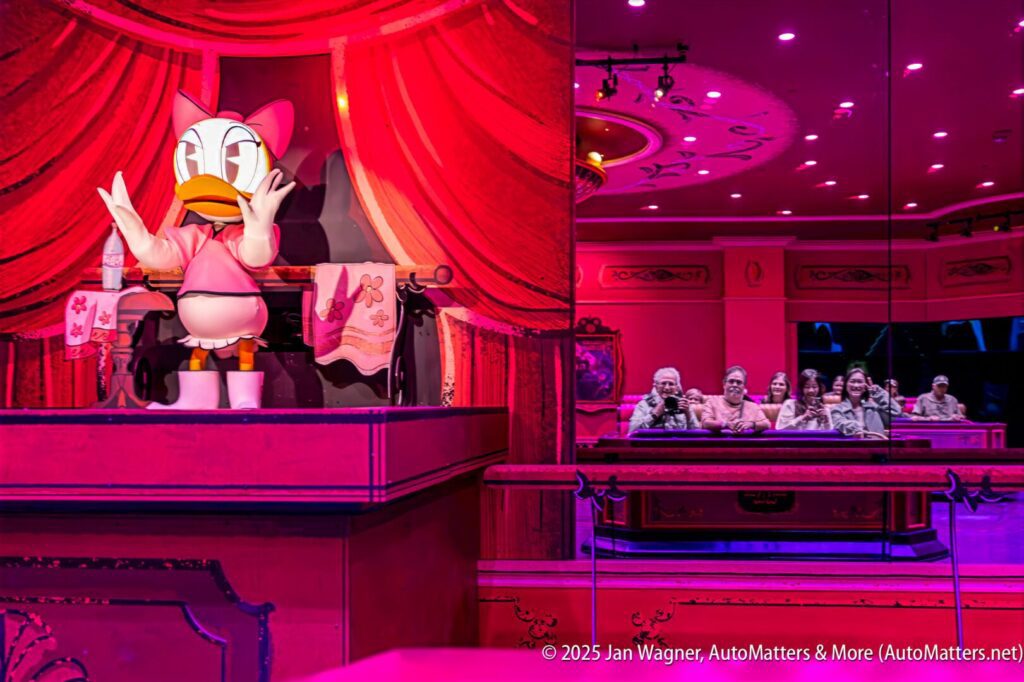 An animatronic Daisy Duck performs on stage while a group of people watch from a ride vehicle in a themed attraction.