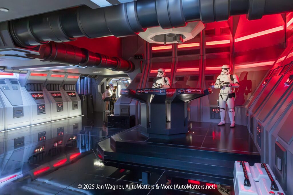A futuristic sci-fi control room with two people in white armor suits standing at consoles, surrounded by red and gray lighting and panels.