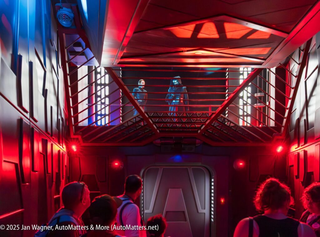 People stand in a dimly lit, futuristic corridor with red and blue lighting, looking up at two costumed characters positioned on a raised platform behind a metal railing.