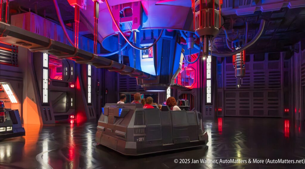 A group of people ride in a trackless vehicle inside a futuristic, sci-fi themed attraction with colorful lighting and industrial details.