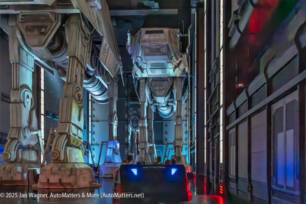Two large AT-AT walker models stand in a themed indoor attraction with visitors seated in a dark ride vehicle below.