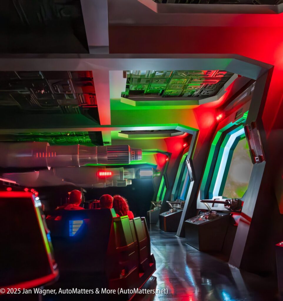 People sit in a themed ride vehicle, surrounded by futuristic decor with red and green lighting, resembling a spaceship interior.