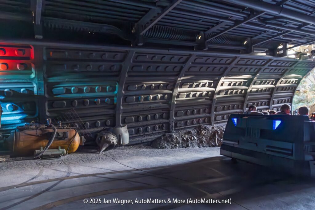 A futuristic vehicle with blue lights drives through a dimly lit, industrial-themed tunnel with machinery and metal walls.