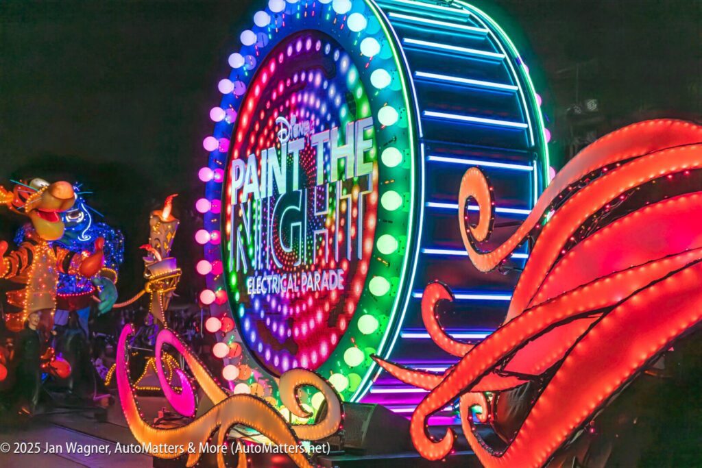A brightly lit float with the words "Paint the Night Electrical Parade" moves through a nighttime parade, surrounded by colorful lights and illuminated decorations.
