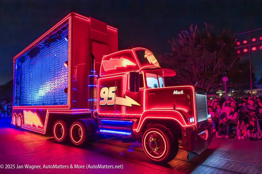 A brightly lit semi-truck float resembling Mack from "Cars," outlined in red neon lights, drives in a nighttime parade with a crowd watching.