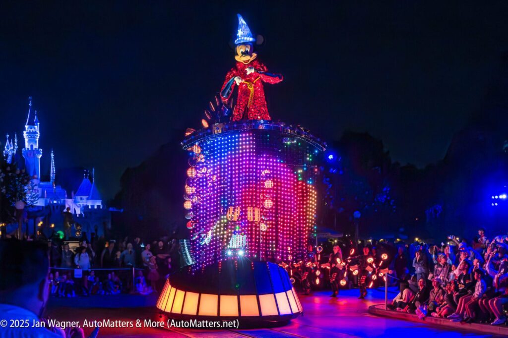 A parade float with a brightly lit Sorcerer Mickey Mouse figure passes by crowds at night in Disneyland, with a castle illuminated in the background.