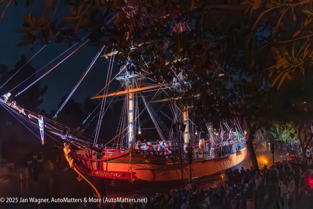 A large, illuminated sailing ship with tall masts is docked at night, surrounded by a crowd of people observing from the shore.