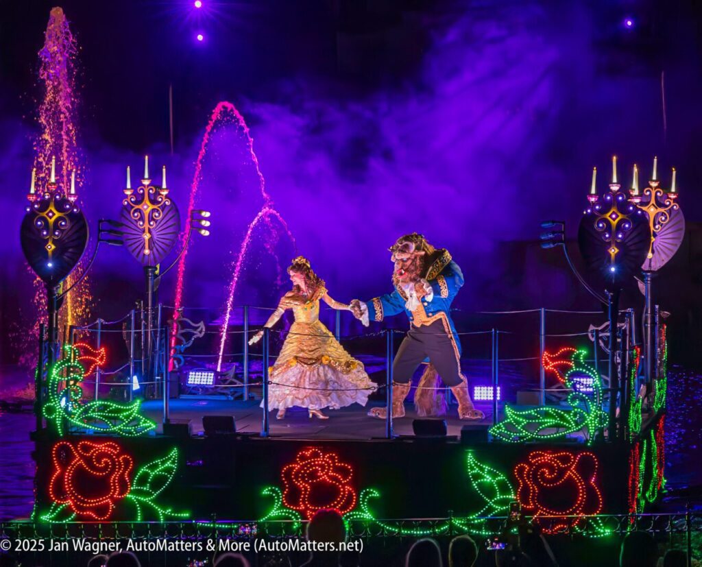 Two performers dressed as Belle and the Beast dance on an illuminated float, surrounded by glowing roses and candelabras, with pink water fountains in the background at night.