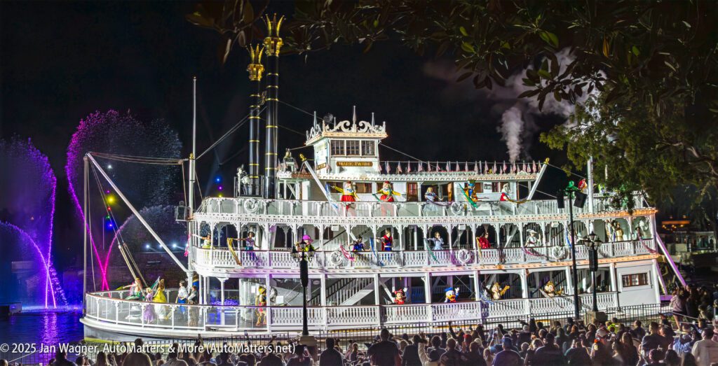 A large riverboat decorated with lights floats on water at night, with performers on board and colorful water fountains in the background.